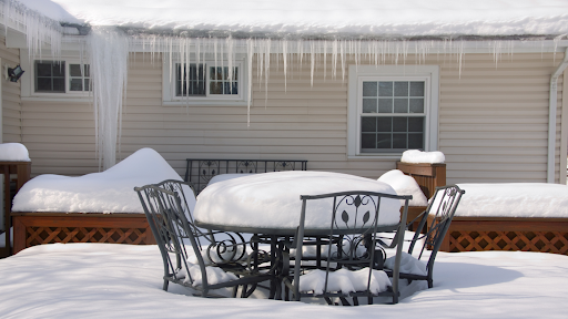 A snow-covered patio with a table and chairs, a hot tub with a closed lid, and a house with icicles hanging from the roof.